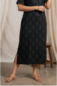 Person wearing a black patterned dress with a wooden stool and beige curtain in the background