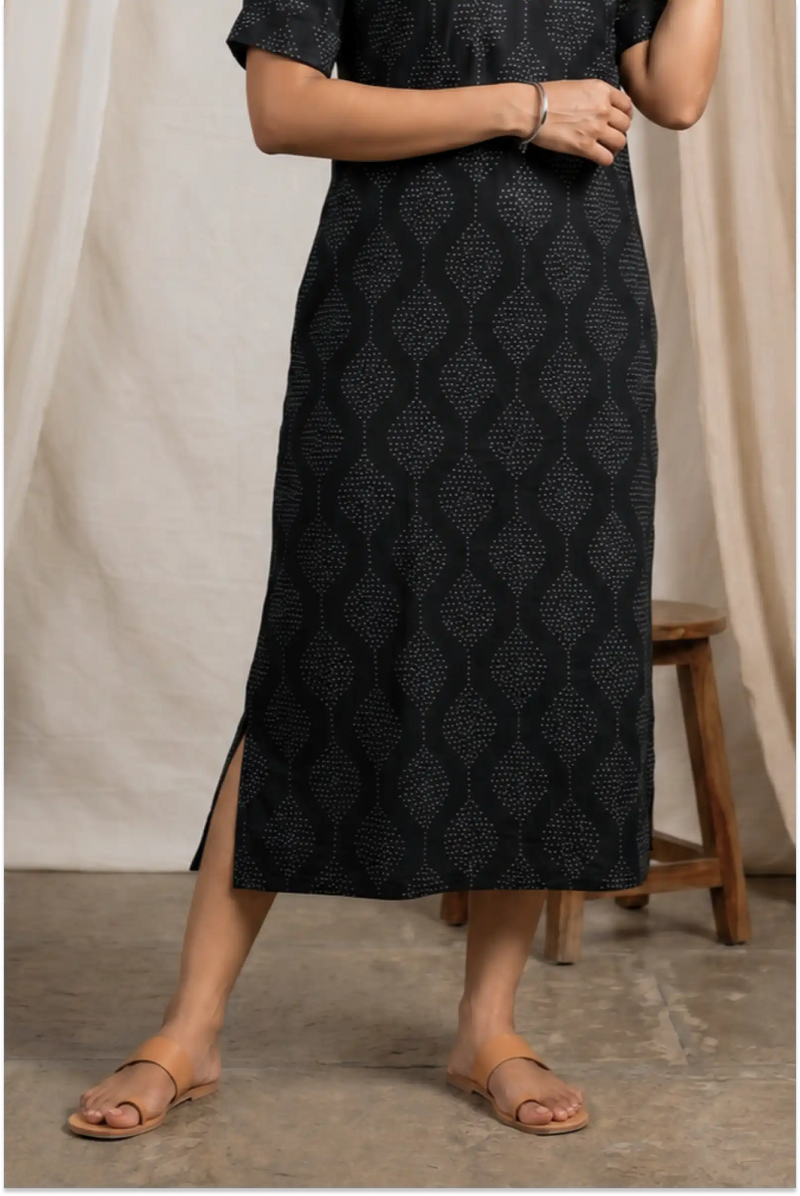 Person wearing a black patterned dress with a wooden stool and beige curtain in the background