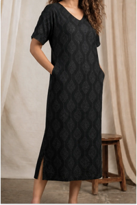 Woman wearing a black patterned dress standing against a beige curtain.